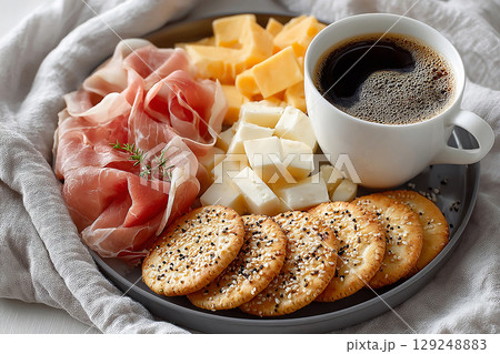 Plate with slices prosciutto, pieces of brie, cheddar with crackers and cup of coffee on a white background. Classic Continental Breakfast. Hotel Breakfast Styles. Plate with slices prosciutto, pieces of brie, cheddar with crackers and cup of coffee on a white background. Classic Continental Breakfast. Hotel Breakfast Styles. 129248883