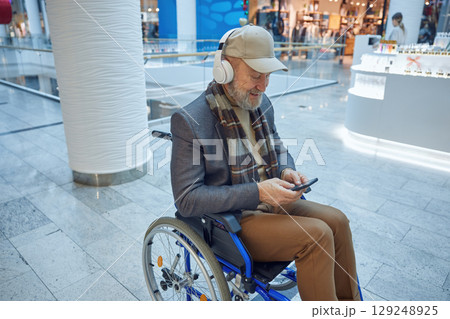 Elderly man comfortably seated in a wheelchair using his smartphone in a modern mall setting 129248925