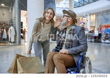 Mature man in a wheelchair is assisted by a woman in a shopping mall 129248928