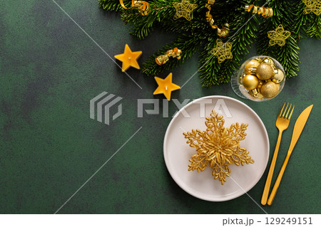 Top view of a Christmas table, featuring an empty plate with cutlery on a green surface. Spruse branches and golden tree decorations set a joyful holiday atmosphere Top view of a Christmas table, featuring an empty plate with cutlery on a green surface. Spruse branches and golden tree decorations set a joyful holiday atmosphere 129249151
