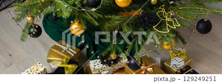 Sophisticated gold and black-toned gift boxes paired with glowing candles and intricate ornaments, nestled under the Christmas tree for a warm and modern holiday celebration, banner 129249251