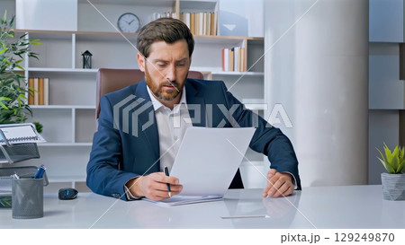 Businessman dressed in a tailored suit and tie, sitting at a modern office desk, intently reading documents while holding a pen, deeply focused on the important paperwork 129249870