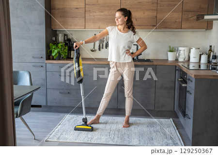 Young woman after vacuuming carpet with wireless vacuum cleaner in kitchen. Household and useful technology concept.  129250367