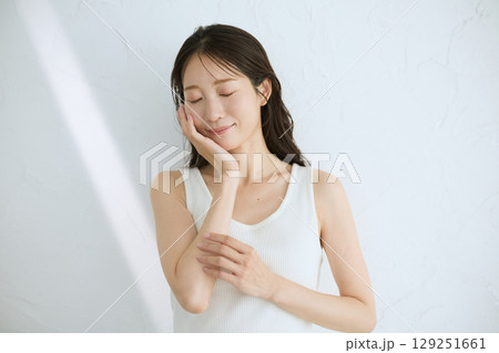 肌に触れてスキンケアする若い女性の笑顔 – 自然光・白背景 129251661