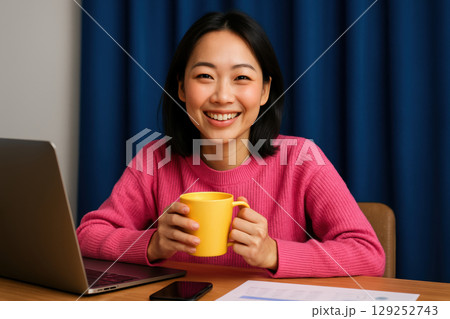 Smiling young woman in pink sweater holding yellow mug while working on laptop at desk with documents and smartphone, AI Generative 129252743