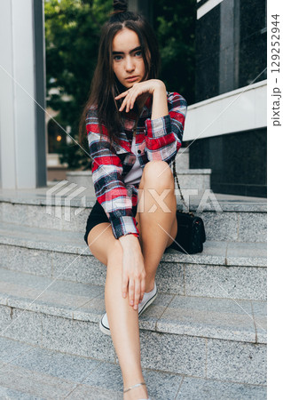 Woman in a plaid shirt and black shorts sits on a set of stairs 129252944