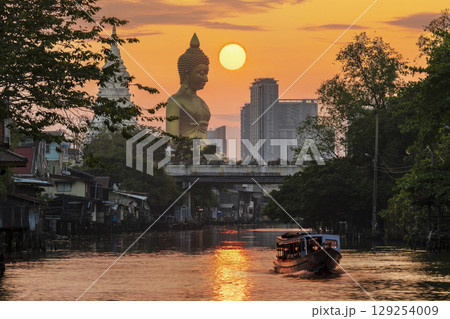 A boat was passing Big Buddha statue (Phra Buddha Dhammakaya Thepmongkhon) in Wat Pak Nam Phasi Charoen temple located by the river during sunset. 129254009