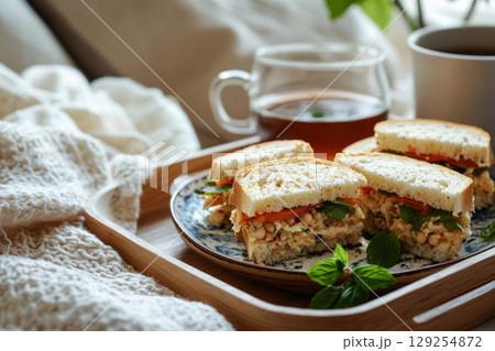Delicious breakfast spread on a wooden platter showcases freshly made sandwiches filled with vegetables and a side of warm tea. Sunlight enhances the inviting atmosphere in the room 129254872