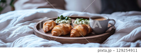 A wooden tray holds three buttery croissants and two small bowls of green dip, accompanied by a steaming cup of coffee, resting on soft bedding, creating a tranquil breakfast setting A wooden tray holds three buttery croissants and two small bowls of green dip, accompanied by a steaming cup of coffee, resting on soft bedding, creating a tranquil breakfast setting 129254911