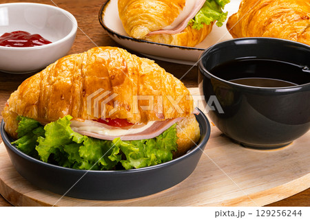 Croissant ham cheese in black ceramic dish and a cup of black coffee on wooden cutting board. 129256244