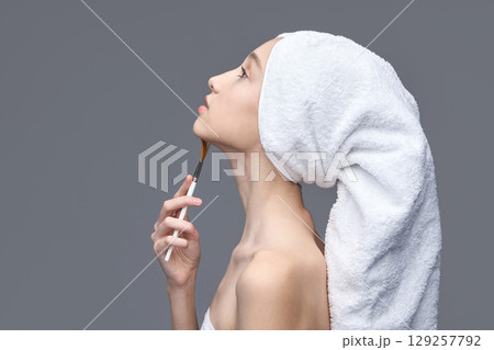 Young caucasian female with towel on head applying makeup brush on neck against Young caucasian female with towel on head applying makeup brush on neck against 129257792