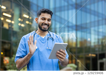An Indian doctor waves during a video call on a tablet outside a modern building. 129259406