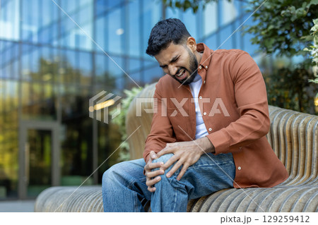 A man clutches his knee in pain, his face contorted with discomfort. He is seated outside near a building with a glass facade. 129259412