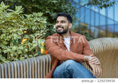 A young, smiling man sits outdoors, enjoying a sunny day. He is wearing casual attire, surrounded by greenery and modern architecture. 129259416