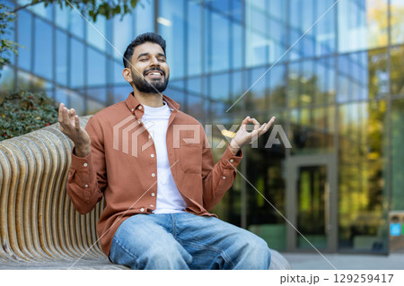A man in a meditative pose outdoors. He sits with his eyes closed, demonstrating mindfulness and relaxation. 129259417
