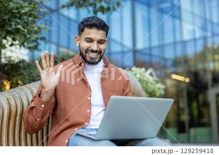 An Indian man smiles, waving during a video call on his laptop outdoors in front of an office building. 129259418