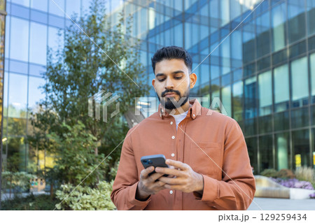 A man using a smartphone outside a modern office building, with a thoughtful expression. 129259434