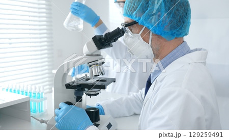 Masked male scientist examining specimen under microscope while laboratory assistant handles glassware in a professional medical investigation at the background. Medicine, science concept 129259491