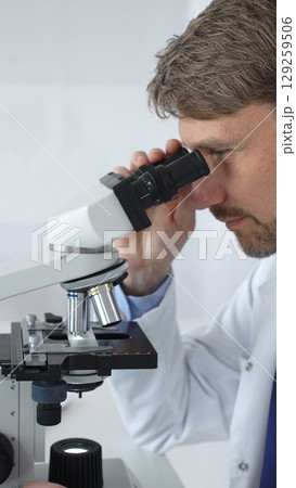 A male scientist looks through a microscope in a brightly lit laboratory while conducting research to develop new drugs and treatments. Medicine and science concept A male scientist looks through a microscope in a brightly lit laboratory while conducting research to develop new drugs and treatments. Medicine and science concept 129259506