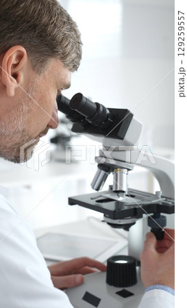 A male scientist looks through a microscope in a brightly lit laboratory while conducting research to develop new drugs and treatments. Medicine and science concept A male scientist looks through a microscope in a brightly lit laboratory while conducting research to develop new drugs and treatments. Medicine and science concept 129259557