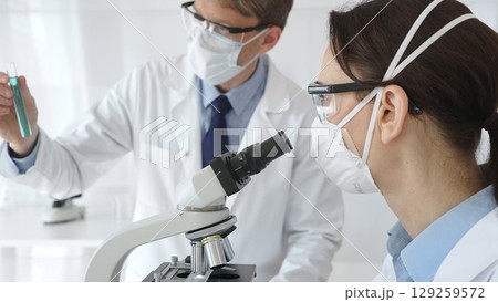 Two scientists in lab coats and protective gear conduct research in a sterile laboratory: one observes through a microscope, and the other holds a test tube with turquoise liquid. Medicine and science 129259572