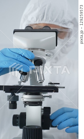 Male scientist virologist wearing hazmat suit, mask, and blue gloves, diligently working at a microscope in a laboratory researching viruses. Medicine and science concept 129259573