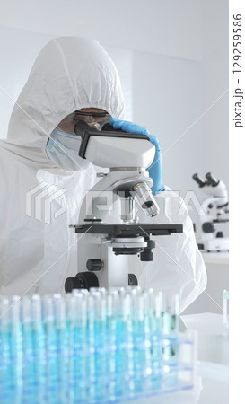 Medical scientist wearing protective suit using microscope while analyzing samples, conducting research in laboratory environment. Close up of test tubes with blue liquid Medical scientist wearing protective suit using microscope while analyzing samples, conducting research in laboratory environment. Close up of test tubes with blue liquid 129259586