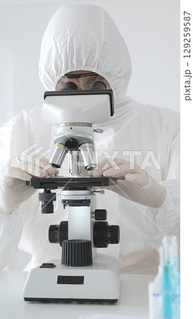 Medical virologist in protective hazmat gear examining viral samples under a microscope while conducting research in a sterile laboratory environment 129259587