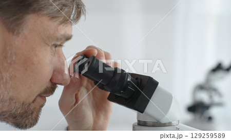Close up of a man scientist carefully observing through a microscope in a brightly lit laboratory, conducting research for developing new medicines and treatments. Medicine and science concept Close up of a man scientist carefully observing through a microscope in a brightly lit laboratory, conducting research for developing new medicines and treatments. Medicine and science concept 129259589