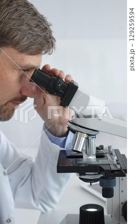 A male scientist looks through a microscope in a brightly lit laboratory while conducting research to develop new drugs and treatments. Medicine and science concept 129259594