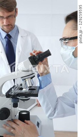 Two scientists, a man and a woman, in lab coats and protective gear, conduct research in a sterile laboratory: one looks through a microscope, and the other holds a test tube with turquoise liquid 129259595