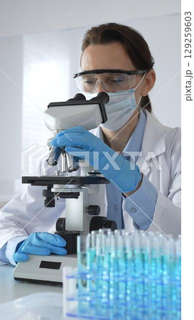 Female scientist examining microscope slide, wearing protective gear in laboratory, researching pharmaceutical breakthrough with blue tinted test tube specimens 129259603