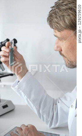 Senior male scientific researcher wearing white lab coat, analyzing blue liquid sample with tablet and microscope during medical investigation. Medicine and science concept Senior male scientific researcher wearing white lab coat, analyzing blue liquid sample with tablet and microscope during medical investigation. Medicine and science concept 129259610