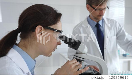 Female scientist wearing protective glasses looking through a microscope during medical research in laboratory with her male colleague working in the background. Medicine and science concept Female scientist wearing protective glasses looking through a microscope during medical research in laboratory with her male colleague working in the background. Medicine and science concept 129259613