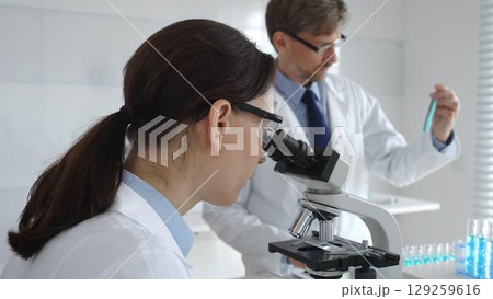 Female scientist wearing protective glasses looking through a microscope during medical research in laboratory with her male colleague working in the background. Medicine and science concept Female scientist wearing protective glasses looking through a microscope during medical research in laboratory with her male colleague working in the background. Medicine and science concept 129259616