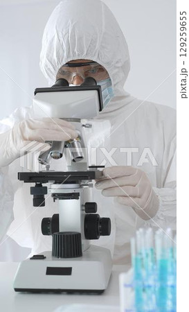 Medical virologist in protective hazmat gear examining viral samples under a microscope while conducting research in a sterile laboratory environment 129259655