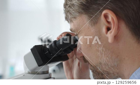 Close up of a man scientist carefully observing through a microscope in a brightly lit laboratory, conducting research for developing new medicines and treatments. Medicine and science concept 129259656