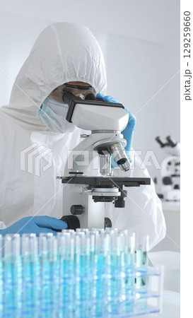 Medical scientist in protective suit using microscope while analyzing samples, conducting research in sterile laboratory environment. Close-up of test tubes with blue liquid 129259660