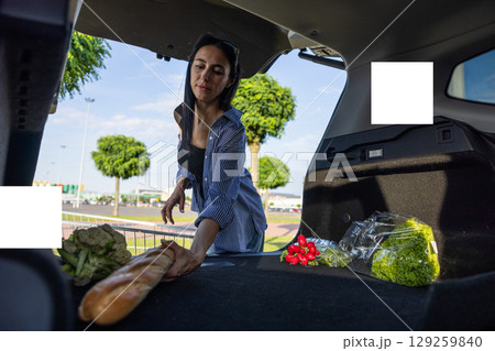 young brunette woman standing in supermarket parking lot next to car with open trunk and transferring groceries from shopping cart into open trunk of car young brunette woman standing in supermarket parking lot next to car with open trunk and transferring groceries from shopping cart into open trunk of car 129259840