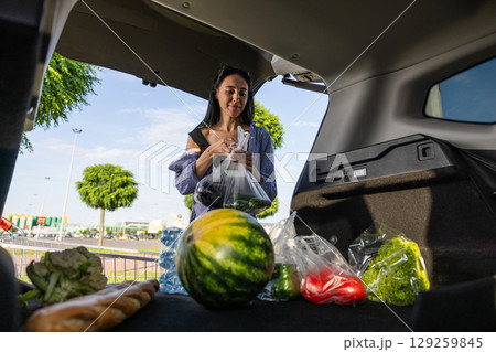 young brunette woman standing in supermarket parking lot next to car with open trunk and transferring groceries from shopping cart into open trunk of car 129259845