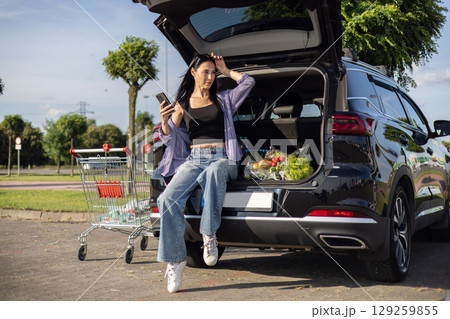 young brunette woman in casual clothes in supermarket parking lot sitting in open car trunk filled with groceries 129259855