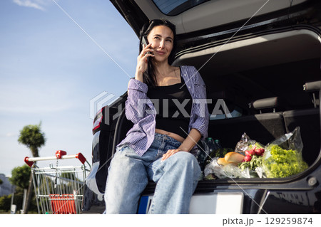 young brunette woman in casual clothes in supermarket parking lot sitting in open car trunk filled with groceries 129259874