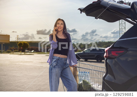 young brunette woman standing in supermarket parking lot with shopping cart next to car with open trunk and talking on phone young brunette woman standing in supermarket parking lot with shopping cart next to car with open trunk and talking on phone 129259881