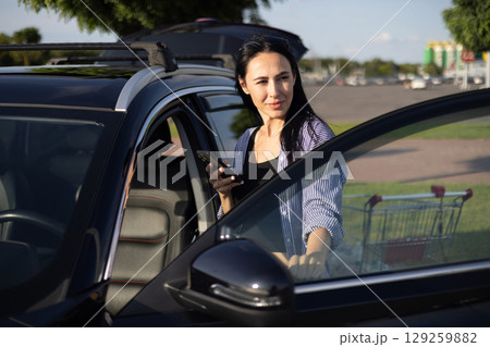young brunette woman in casual clothes in supermarket parking lot standing by car with open door and holding phone in hands young brunette woman in casual clothes in supermarket parking lot standing by car with open door and holding phone in hands 129259882