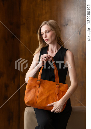 A female model with blonde wavy hair is elegantly posed seated, while holding a vibrant amber leather tote bag against her all-black outfit, creating a sophisticated fashion portrait 129260106
