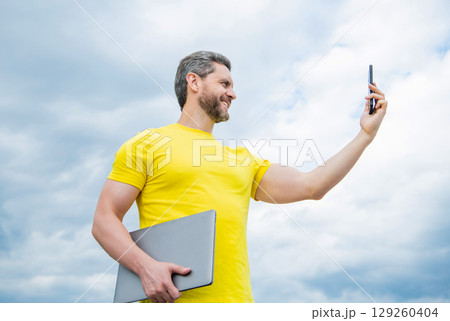 man with laptop having video call on smartphone on sky background 129260404