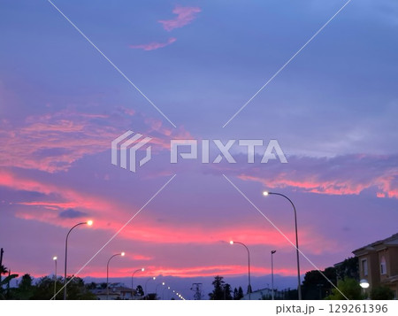 Vibrant pink clouds fill the sky as the sun sets over a quiet city street. Streetlights illuminate the scene, creating an enchanting atmosphere at dusk. 129261396