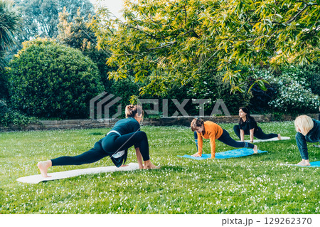 Female yoga class in park. Group of diverse women doing stretching pose exercising together with instructor on green grass lawn. Sport activity for health, wellbeing, mental health. Selective focus 129262370