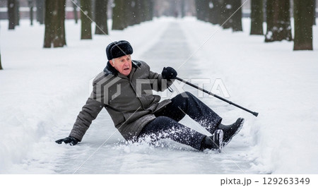 An elderly man loses his balance and slips on ice while walking in a snowy park. He is visibly startled and trying to stabilize himself using a cane amidst the winter scenery. 129263349
