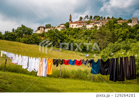 Scenic Hilltop Town Of Stanjel In Slovenia: Traditional Stone Houses And Historic Church Tower Rising Above Lush Greenery Under A Dramatic Cloudy Sky In The Karst Region 129264281